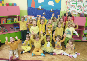 Dzieci z grupy drugiej, w żółtych strojach śmieją się unosząc ręce do góry